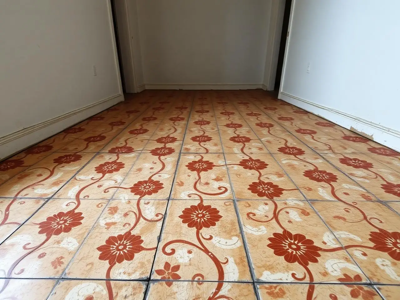 Pièce vide vue au ras du sol : anciens carreaux à motifs floraux rouge brique et crème, émail usé et joints noircis.