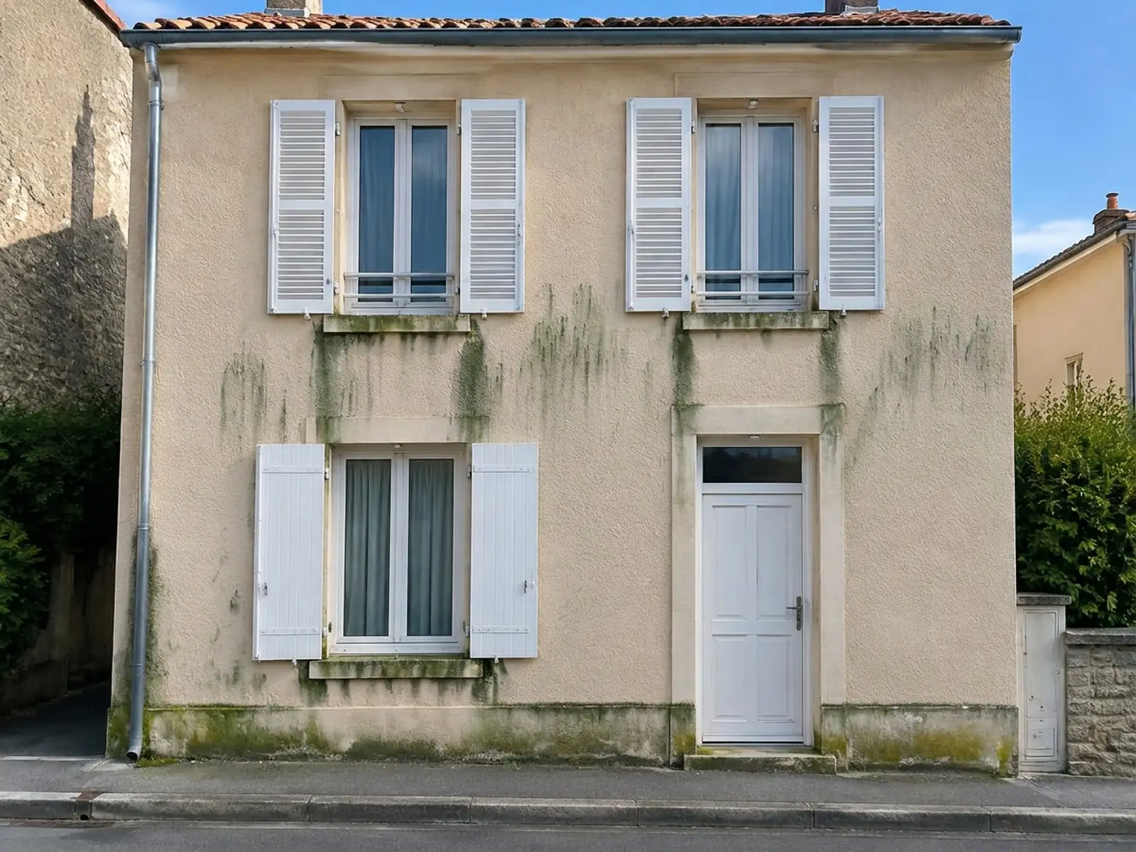 Façade enduite d’une maison de ville à volets blancs : coulures vertes d’algues sous les appuis de fenêtre, pied de mur verdi et encrassé.